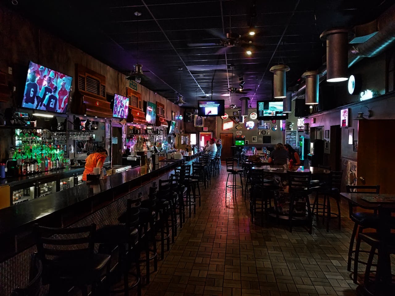A dimly lit modern bar interior featuring a long counter and multiple screens, creating a cozy urban atmosphere.